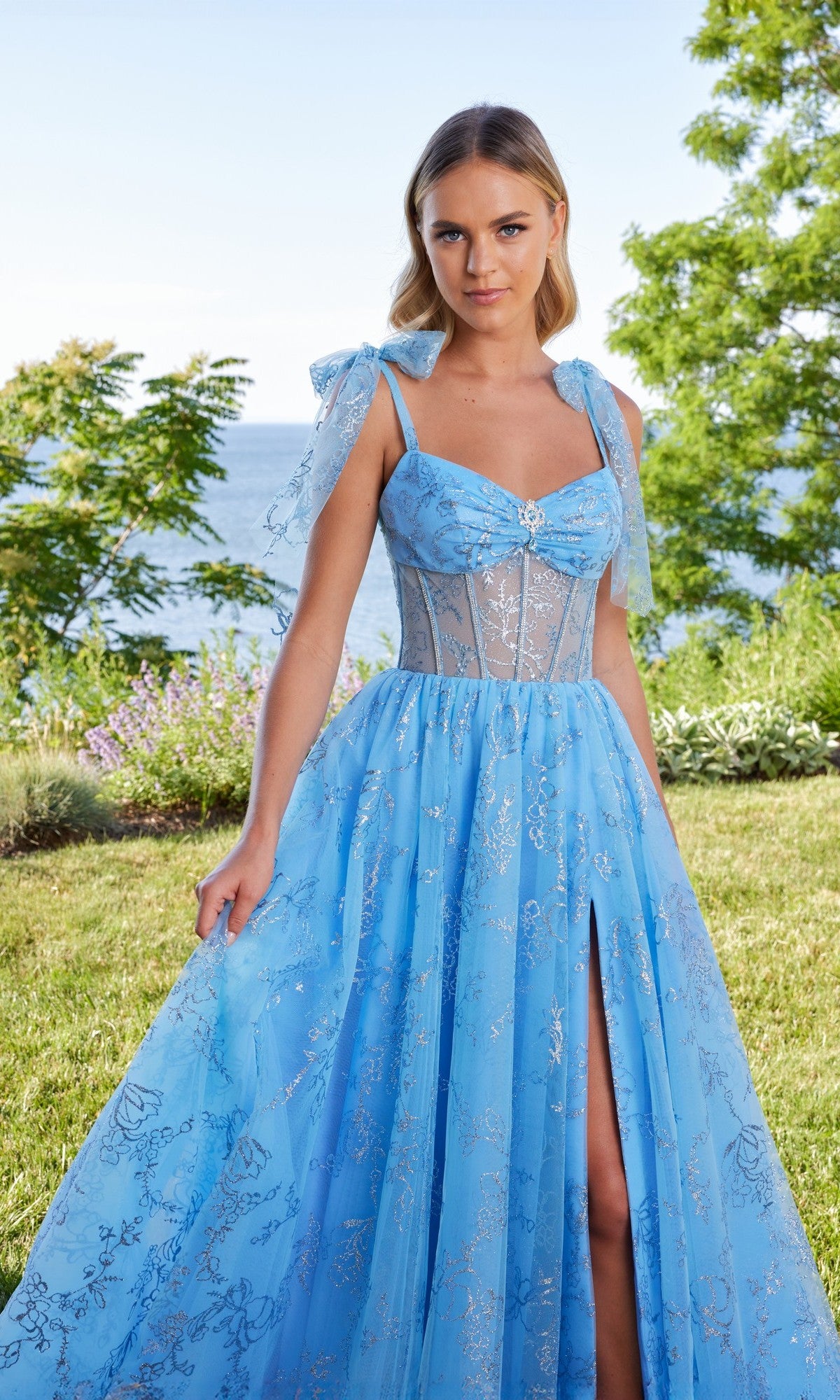 A woman in the Morilee 49185 long prom dress, featuring a light-blue sheer corset bodice and elegant a-line tulle skirt, stands outdoors on grass, surrounded by lush greenery with water in the background.